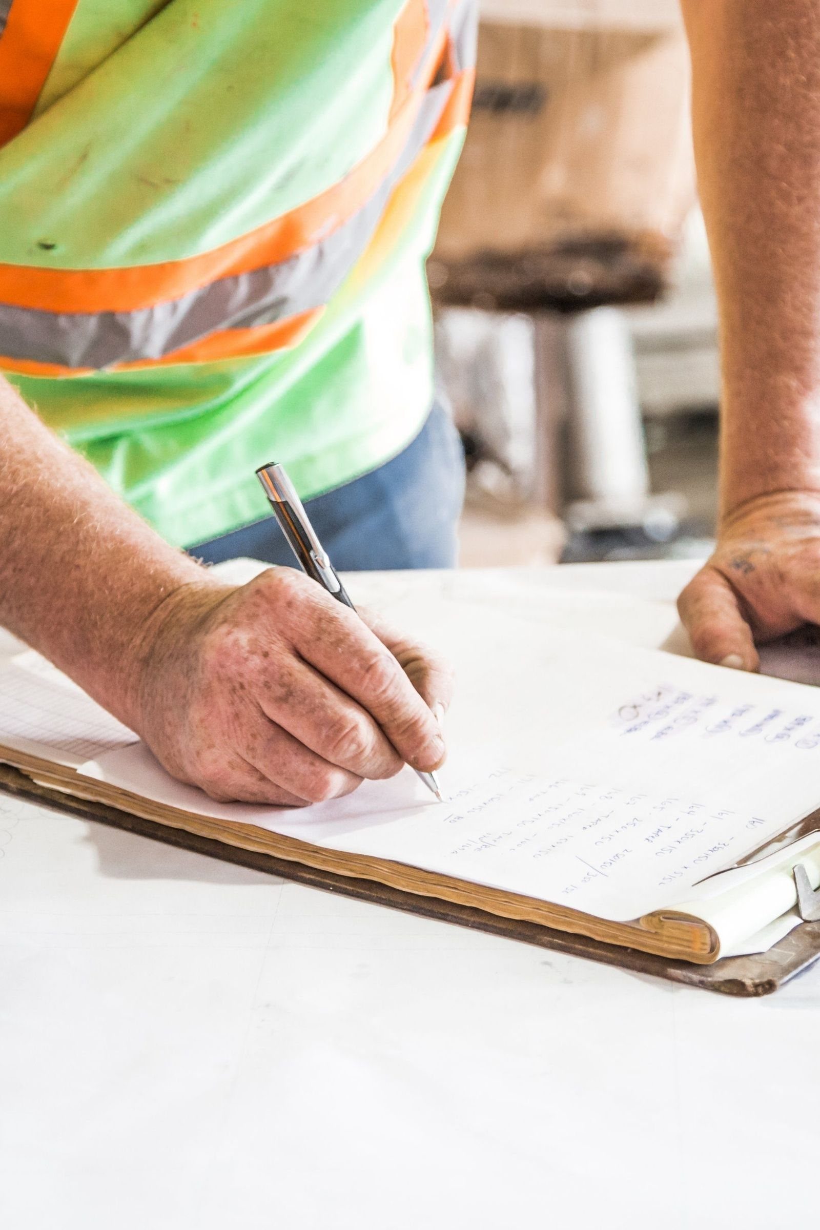 A construction worker in a hi-vis vest writing on a clipboard at a job site, both hands and the handwritten paperwork in focus.